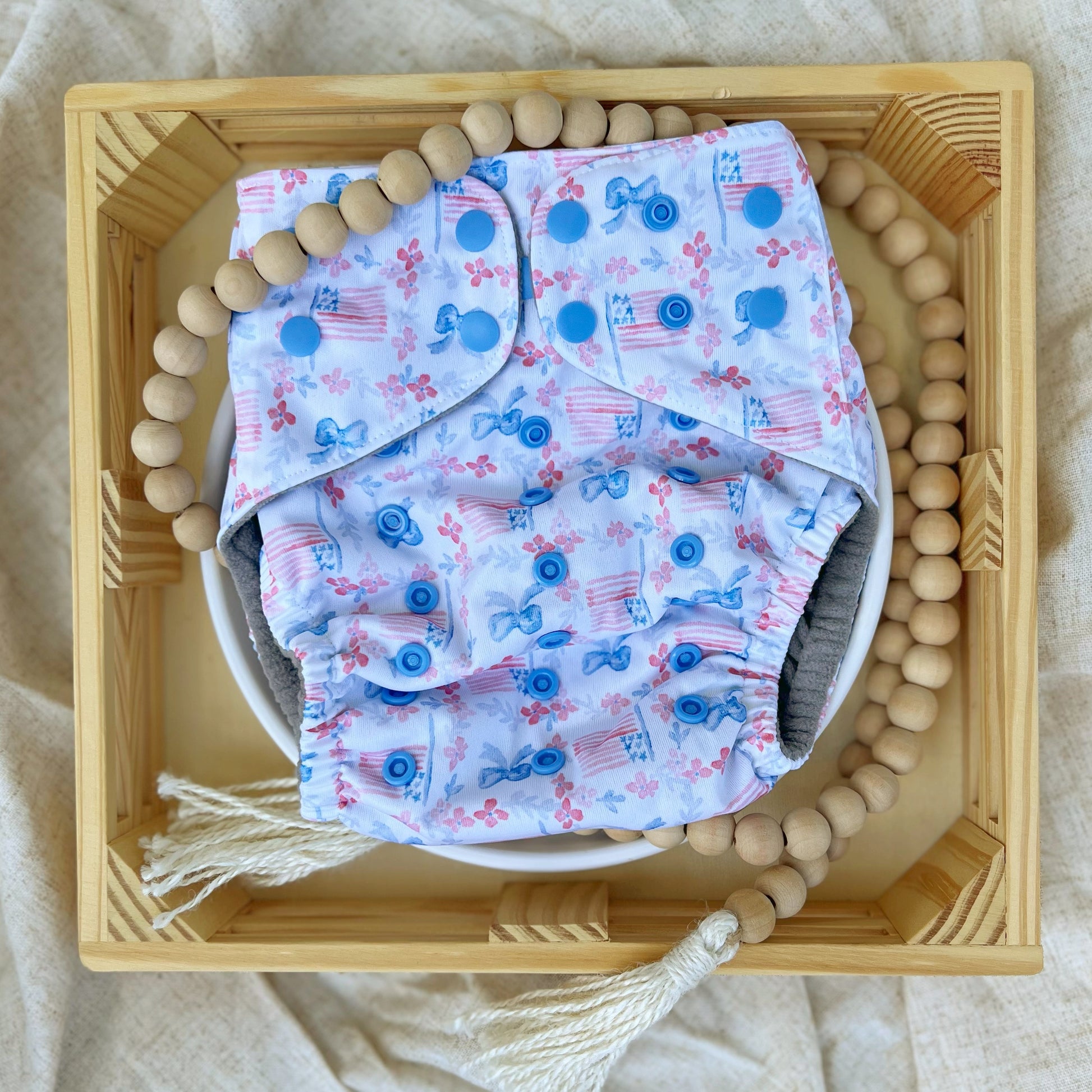 American Beauty. Cloth diaper print with red flowers and blue ferns together running in lines perpendicular to each other, making squares. Blue bowties and American flags fill the squares. White background