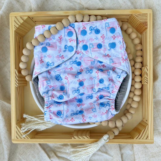 American Beauty. Cloth diaper print with red flowers and blue ferns together running in lines perpendicular to each other, making squares. Blue bowties and American flags fill the squares. White background