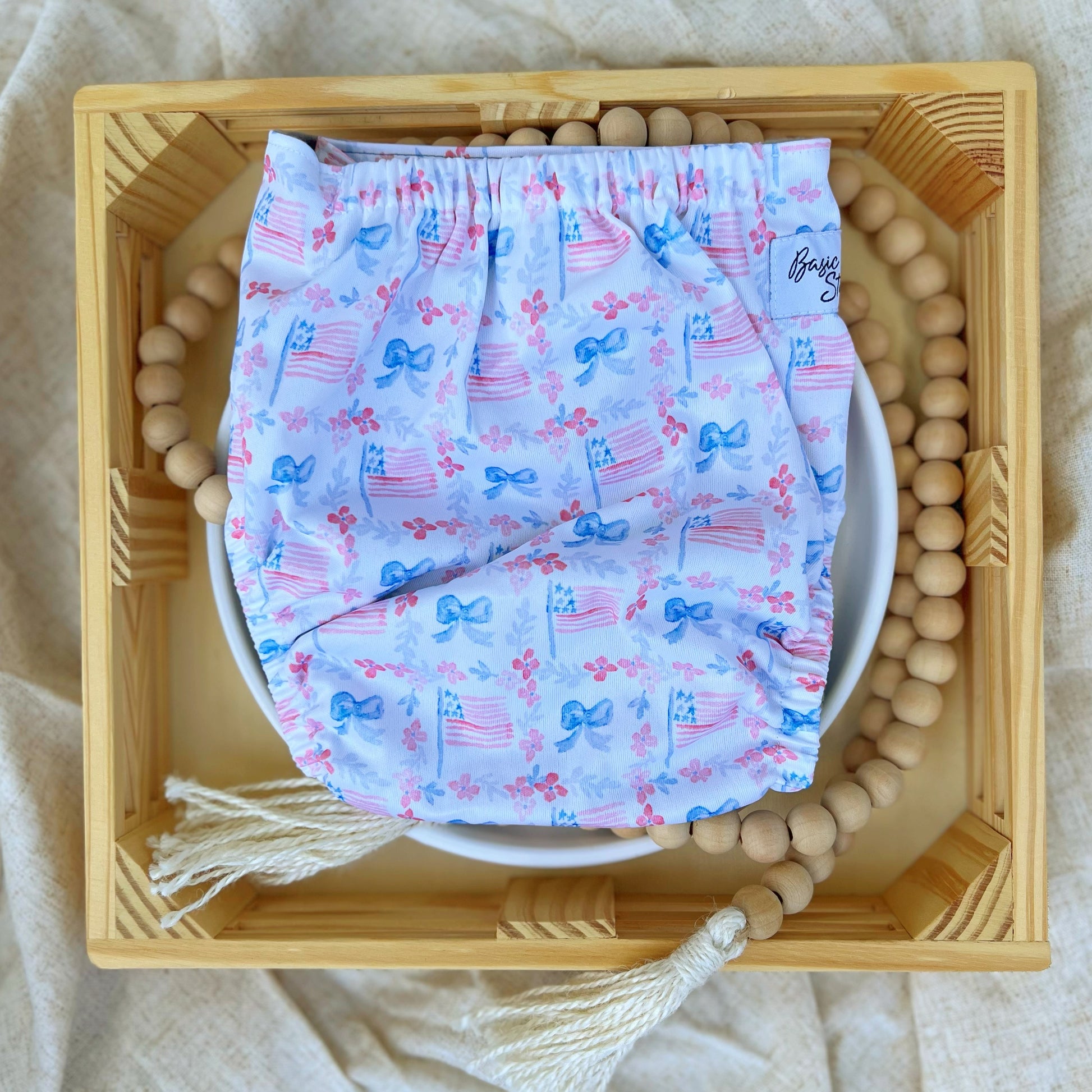 American Beauty. Cloth diaper print with red flowers and blue ferns together running in lines perpendicular to each other, making squares. Blue bowties and American flags fill the squares. White background